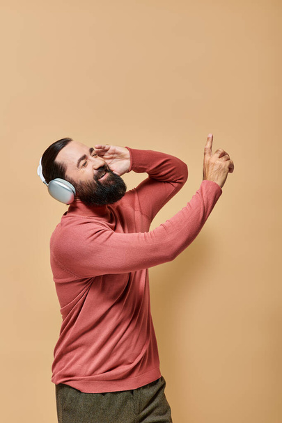 bearded and cheerful man in turtleneck jumper listening music in wireless headphones, beige backdrop - Photo, Image