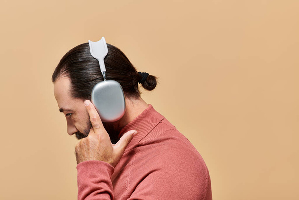 side view, bearded man in turtleneck jumper listening music in wireless headphones on beige - Zdjęcie, obraz