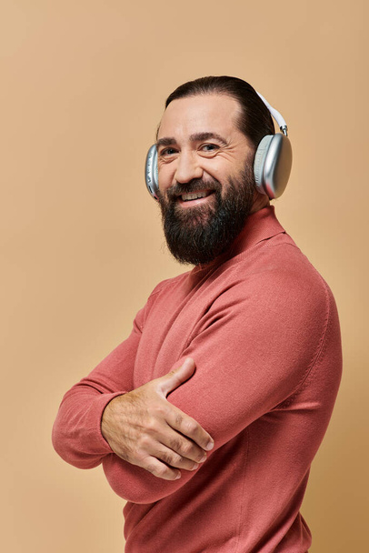 позитивный и бородатый мужчина в водолазке джампер слушать музыку в беспроводных наушниках, бежевый фон - Фото, изображение