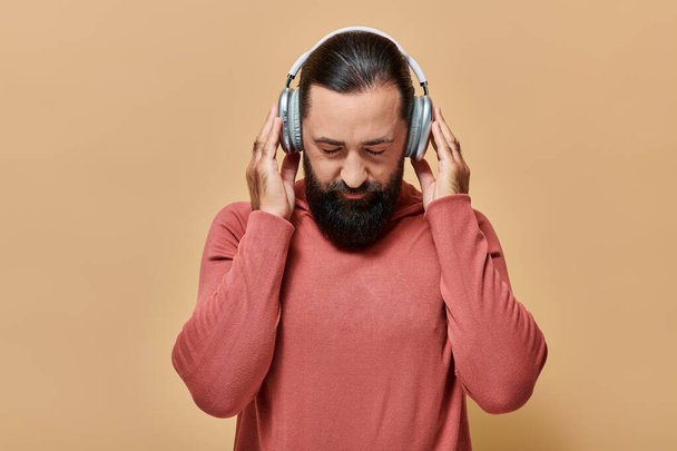 hombre barbudo guapo en jersey de cuello alto escuchando música en auriculares inalámbricos sobre fondo beige - Foto, Imagen