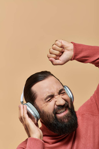 hombre barbudo emocionado en jersey de cuello alto escuchando música en auriculares inalámbricos, fondo beige - Foto, Imagen