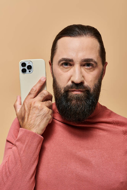 retrato de hombre barbudo de buen aspecto en jersey de cuello alto sosteniendo teléfono inteligente sobre fondo beige - Foto, Imagen
