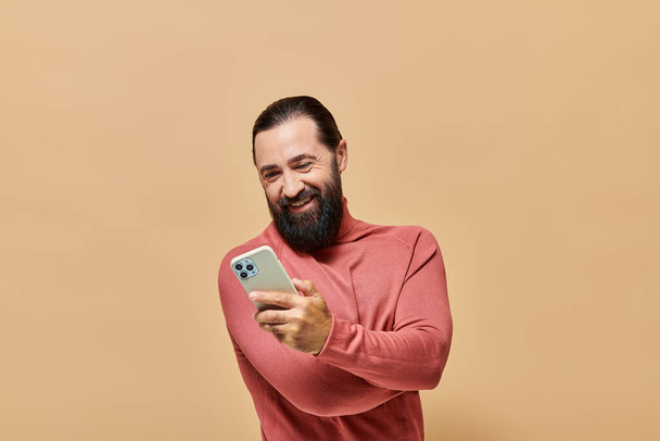portrait of cheerful bearded man in turtleneck jumper holding smartphone on beige background - Photo, Image