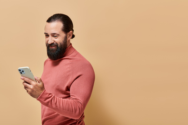 ベージュの背景にスマートフォンを保持するタートルネックジャンパーの陽気なひげ付き男の肖像画 - 写真・画像