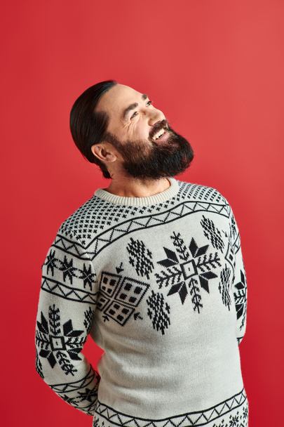 szczęśliwy brodaty mężczyzna w zimowym swetrze z ozdobą uśmiechnięty na czerwonym tle, Wesołych Świąt - Zdjęcie, obraz