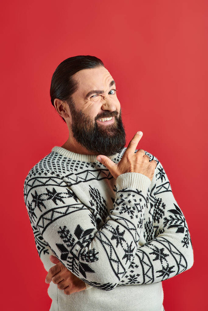 cheerful bearded man in winter sweater with ornament pointing on red backdrop, Merry Christmas - Photo, Image