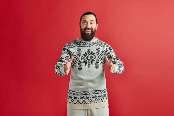 hombre barbudo positivo en suéter de Navidad con adorno gesto sobre fondo rojo, vacaciones de invierno - Foto, Imagen