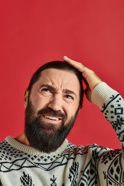 bearded man in Christmas sweater with ornament having headache and touching head on red background - Photo, Image