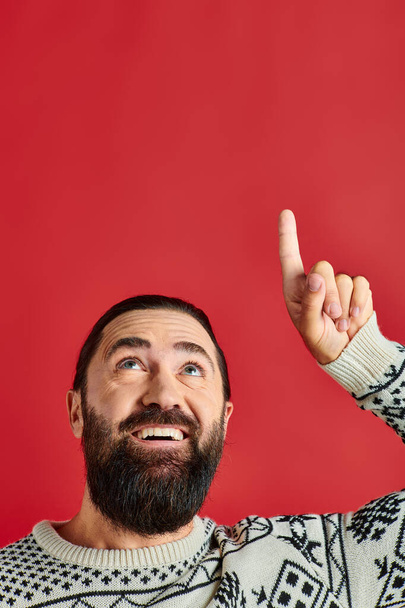 happy bearded man in Christmas sweater with ornament pointing up on red backdrop, winter holidays - Photo, Image