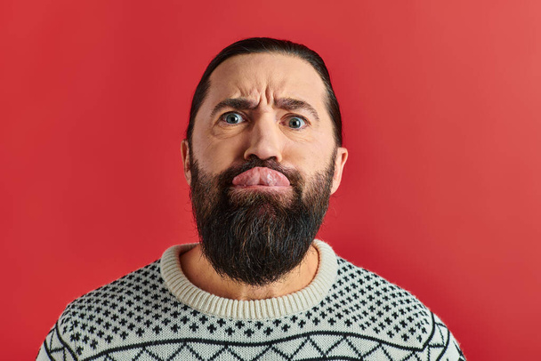 bearded man in winter sweater with ornament sticking tongue out on red backdrop, Christmas - Photo, Image