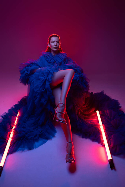 Lenyűgöző modell egy pazar kék karneváli ruhában, elegánsan pózol a neon fények meleg fényénél egy stúdióban.. - Fotó, kép