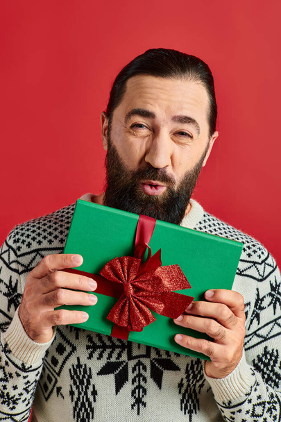 赤い背景にクリスマスプレゼントを持つ冬のセーターのひげ付き男からのエアキス - 写真・画像