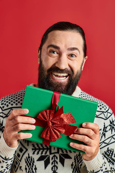 excited bearded man in winter sweater with ornament holding Christmas present on red background - Photo, Image