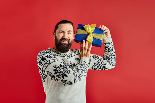 displeased bearded man in winter sweater with ornament holding Christmas present on red background - Photo, Image