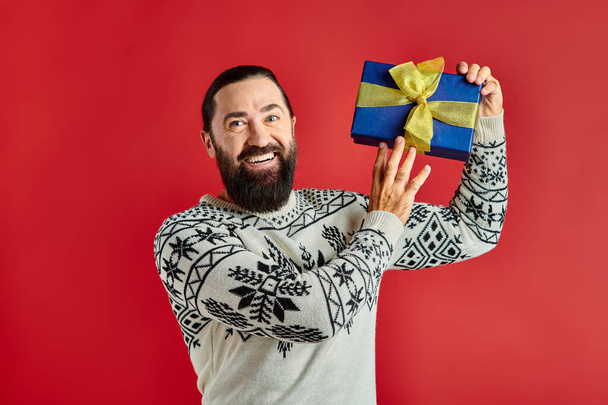 pleased bearded man in winter sweater with ornament holding Christmas present on red background - Photo, Image