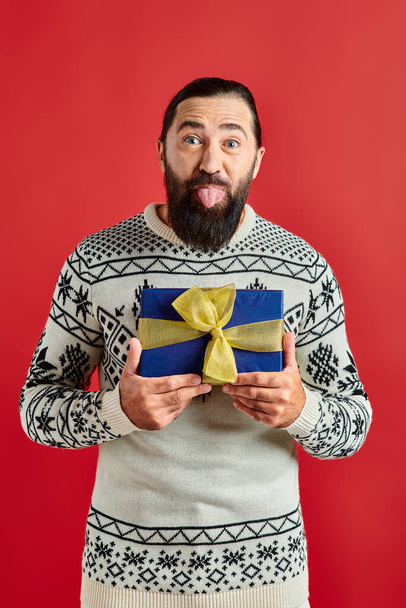 funny bearded man in winter sweater with ornament holding Christmas gift and sticking tongue on red - Photo, Image