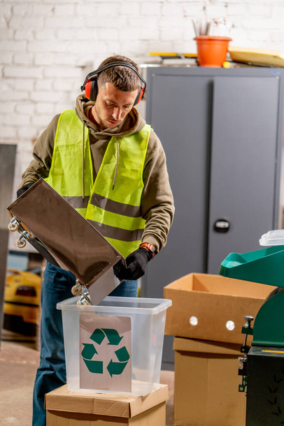 Un empleado de la planta de reciclaje de residuos en uniforme y auriculares vierte tapas de plástico trituradas en un contenedor para reciclar - Foto, Imagen