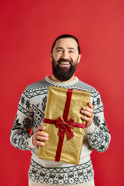 bearded joyful man in winter sweater with ornament holding Christmas present on red background - Photo, Image