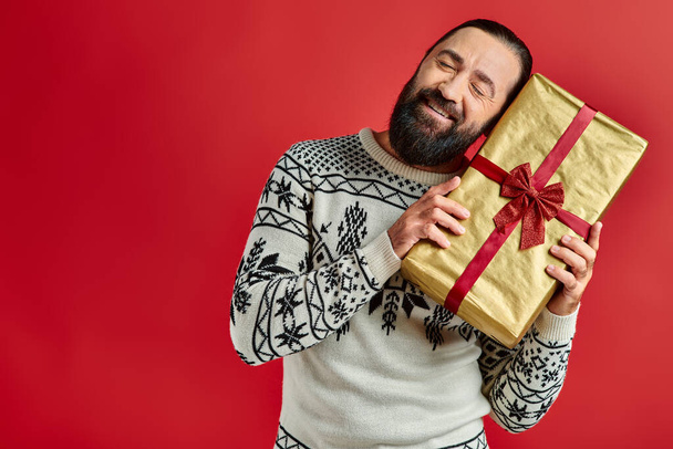 bearded joyful man in winter sweater with ornament holding Christmas present on red background - Photo, Image