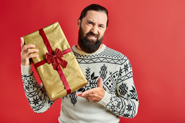 missbilligte bärtige Mann im Winterpullover mit Ornament hält Weihnachtsgeschenk auf rotem Hintergrund - Foto, Bild