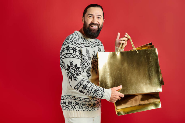 χαρούμενος γενειοφόρος άνδρας σε πουλόβερ με στολίδι κρατώντας τσάντες ψώνια σε κόκκινο φόντο, δώρο Χριστουγέννων - Φωτογραφία, εικόνα