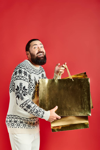 glücklicher bärtiger Mann im Pullover mit Ornament, der Einkaufstüten auf rotem Hintergrund hält, Weihnachtsgeschenk - Foto, Bild