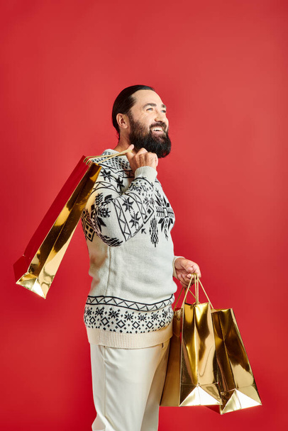 szczęśliwy brodaty mężczyzna w swetrze z ozdobą trzymając torby na zakupy na czerwonym tle, prezent świąteczny - Zdjęcie, obraz