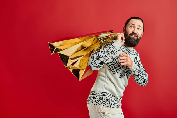 joyful bearded man in sweater with ornament holding shopping bags on red backdrop, Christmas present - Photo, Image