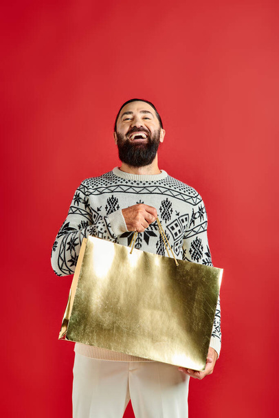smiling bearded man in sweater with ornament holding shopping bags on red backdrop, Christmas time - Photo, Image