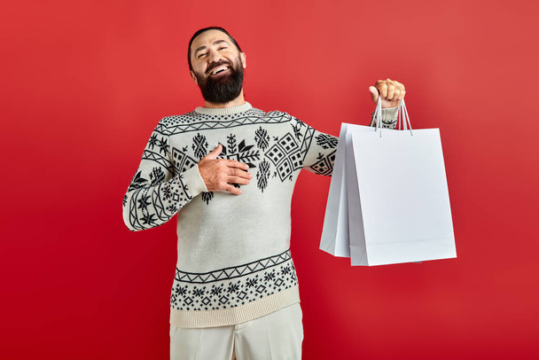 positiver bärtiger Mann im Weihnachtspulli mit Einkaufstüten vor rotem Hintergrund, Feiertagsstimmung - Foto, Bild