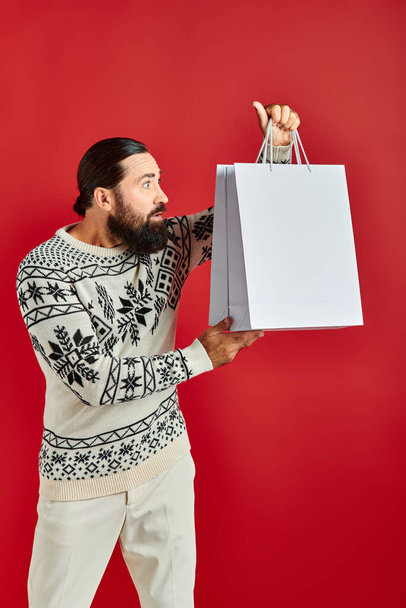 nieuwsgierige man met baard in kersttrui met boodschappentassen op rode achtergrond, feestvreugde - Foto, afbeelding