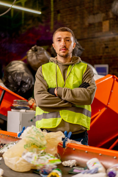 Portret van een serieuze man in een beschermvest met zijn armen voor hem gekruist en kijkend naar de camera in een afvalfabriek - Foto, afbeelding