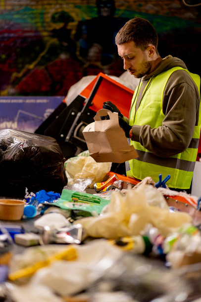 Trieuse masculine ciblée avec gilet de protection et gants décharge les déchets d'un sac poubelle sur une ligne de tri des ordures - Photo, image