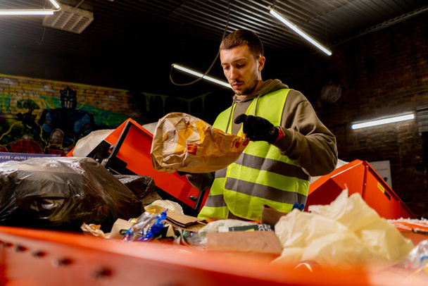 Ένας υπάλληλος με στολή και γάντια σε ένα σταθμό ανακύκλωσης αποβλήτων είδη και ταξινομεί σκουπίδια σε μια γραμμή διαλογής - Φωτογραφία, εικόνα