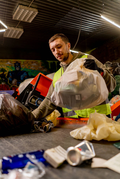 Koruyucu yelek ve eldivenlerle odaklanmış erkek ayırıcı çöp torbasından atıklarını bir çöp sıralama hattına boşaltıyor. - Fotoğraf, Görsel