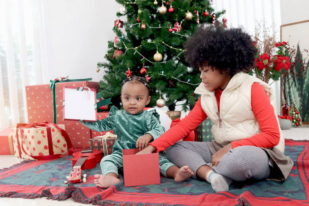 African child sister celebrate winter holiday together, little cute girl kid and curly hair sibling feel excited about Christmas present gift box under beautiful Christmas tree, happy childhood family - Photo, Image