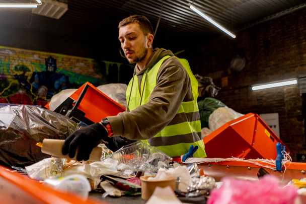 Pracownik w mundurze i rękawiczki w zakładzie recyklingu odpadów sortuje i sortuje śmieci na linii sortującej - Zdjęcie, obraz