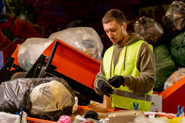 Vista lateral de um homem de uniforme trabalhando em uma linha de distribuição de lixo e classificando resíduos em recipientes especiais - Foto, Imagem