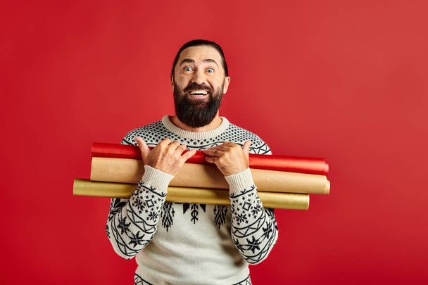Noel süveteri giymiş mutlu sakallı bir adam elinde kırmızı arka planda ambalaj kağıdı, tatil satışları - Fotoğraf, Görsel