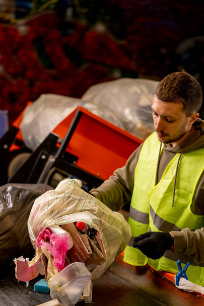Keskittynyt mies lajittelija, jolla on suojaava liivi ja käsineet, purkaa roskapussin jätteet roskien lajitteluriville - Valokuva, kuva