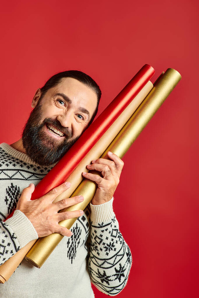 szczęśliwy brodaty mężczyzna w zimowym swetrze trzymając papier pakowy na czerwonym tle, Wesołych Świąt - Zdjęcie, obraz