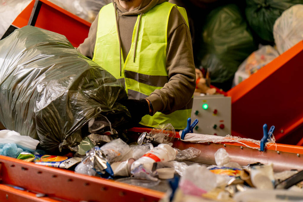 Koruyucu yelek ve eldivenlerle odaklanmış erkek ayırıcı çöp torbasından atıklarını bir çöp sıralama hattına boşaltıyor. - Fotoğraf, Görsel