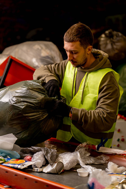 保護ベストと手袋付きの集中男性ソーターは,ゴミの分類ラインにゴミ袋から廃棄物を荷を下ろします - 写真・画像