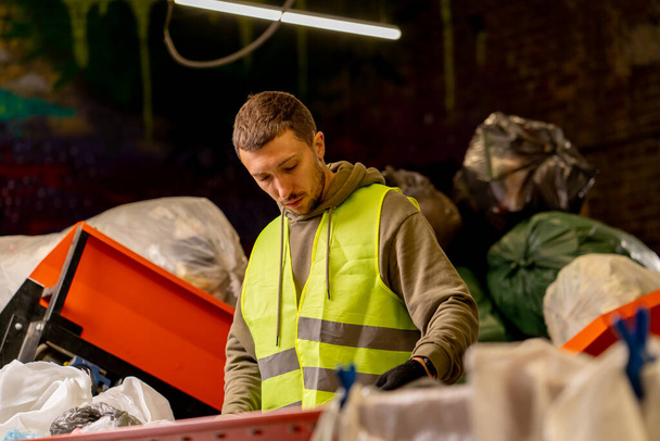 Zijaanzicht van een sorteerder in handschoenen en een beschermvest die afval sorteert op een speciale sorteerlijn tijdens het werken bij een afvalverwerkingsstation - Foto, afbeelding