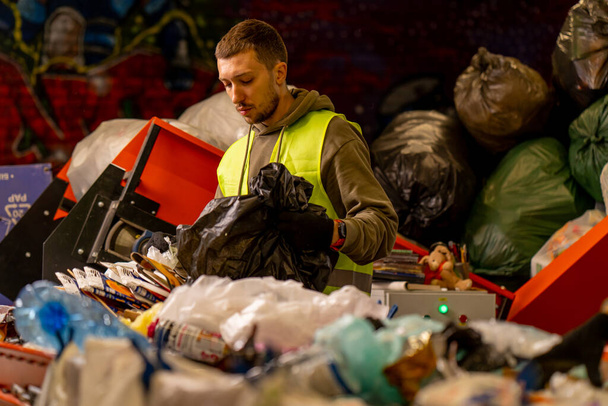 Gerichte mannelijke sorteerder met een beschermvest en handschoenen laadt afval uit een vuilniszak op een afvalsorteerlijn - Foto, afbeelding