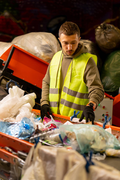 Вид сбоку сортировщика в перчатках и защитного жилета, который сортирует мусор на специальной линии сортировки во время работы на станции утилизации отходов - Фото, изображение