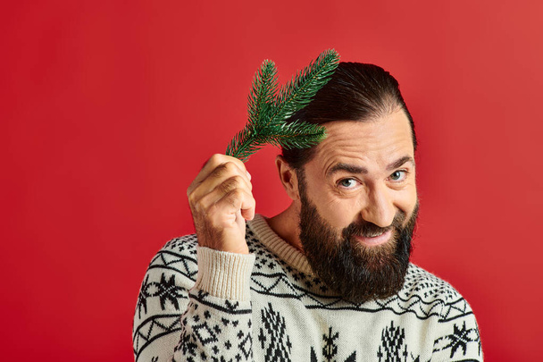 happy bearded man in winter sweater holding branch of pine tree on red backdrop, Merry Christmas - Photo, Image