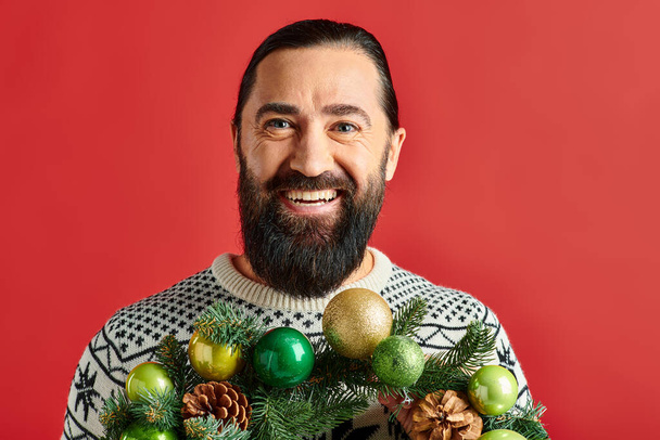 joyful bearded man in winter sweater holding decorated Christmas wreath with baubles on red backdrop - Photo, Image