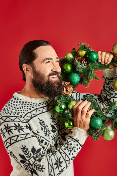 Kış süveteri giymiş mutlu sakallı bir adam elinde süslü bir çelenkle kırmızı arka planda Noel süsleri taşıyor. - Fotoğraf, Görsel