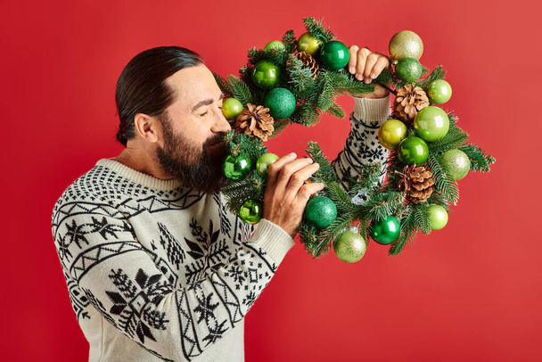 happy bearded man in winter sweater holding decorated wreath with baubles on red backdrop, Christmas - Photo, Image